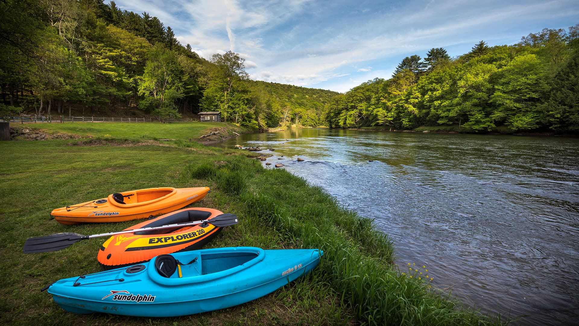 Three colorful kayaks are lined up on the grassy bank of a serene river, surrounded by lush greenery.