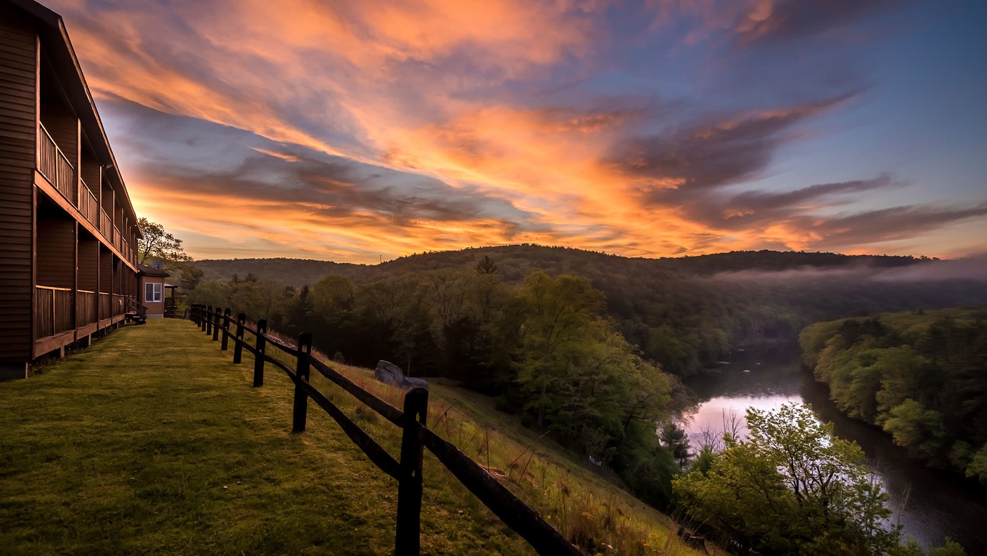 A serene landscape featuring a river, lush hills, and a vibrant sunset, with a wooden building in the foreground.