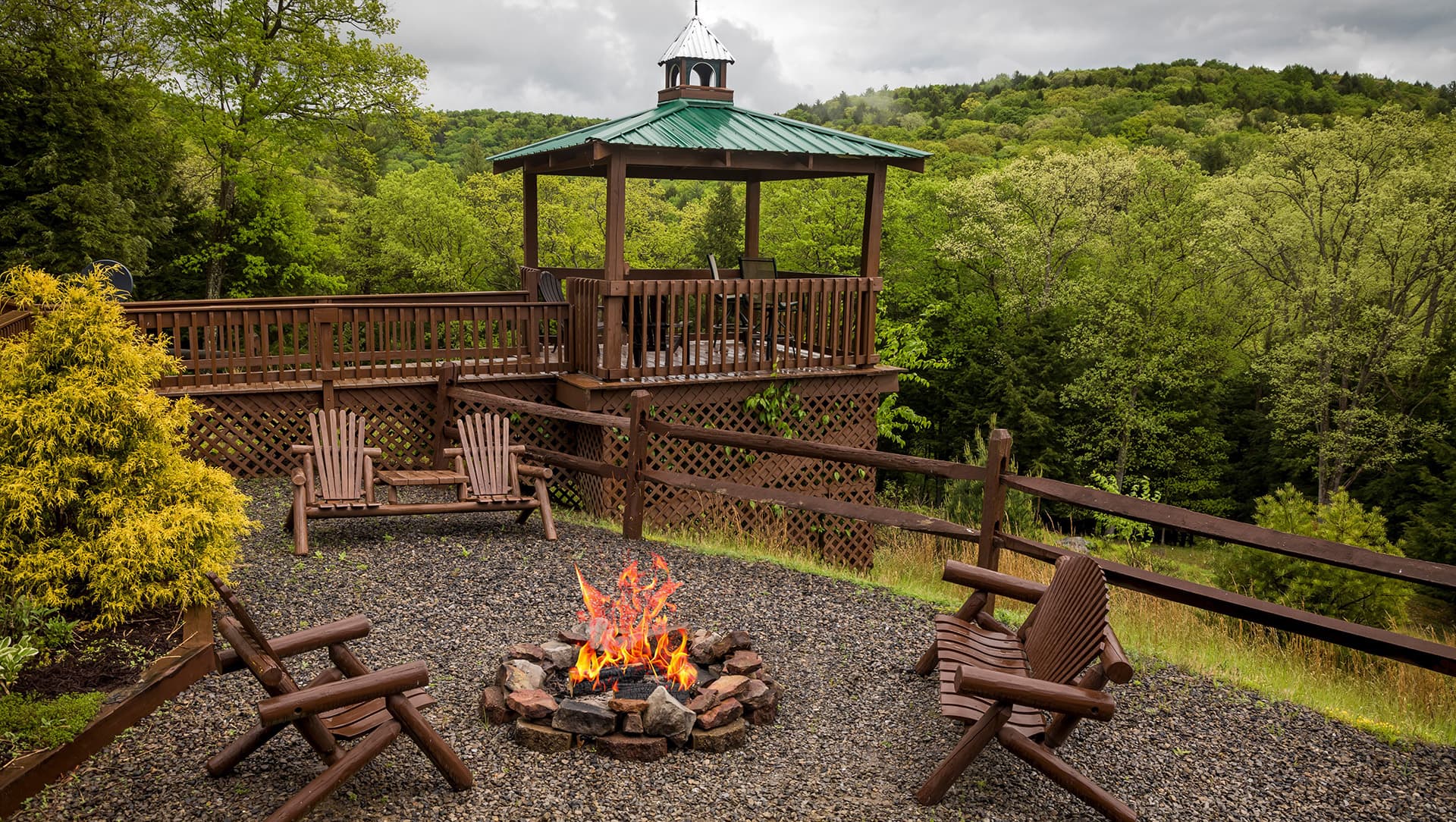 A cozy fire pit with wooden chairs is surrounded by lush greenery and a gazebo in the background.